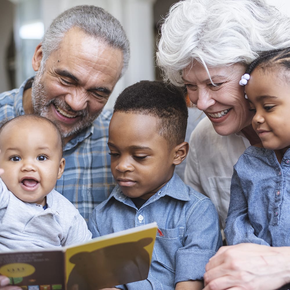 A retired senior couple support family by babysitting. Three young children sit on their grandparents' laps. The group is reading a book together.